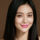 A woman with long straight black hair and pink lipstick smiles at the camera against a plain brown background.