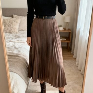 Person taking a mirror selfie wearing a black turtleneck, brown pleated midi skirt with a black belt, and pointed brown ankle boots in a bedroom.