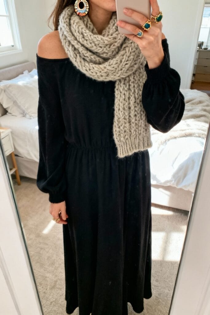 Person wearing a long black off-shoulder dress, chunky knit scarf, large statement earrings, and multiple rings, standing in a bedroom and taking a mirror selfie.