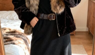 Person taking a mirror selfie, wearing a black dress, brown belt, and a black velvet jacket with a large fur collar in a bedroom.
