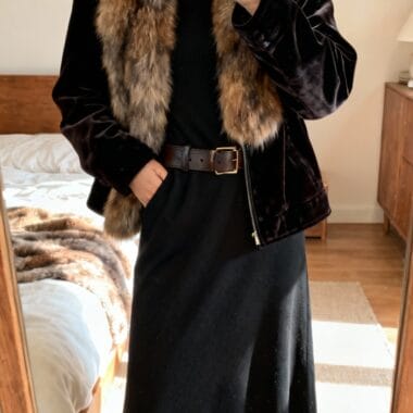 Person taking a mirror selfie, wearing a black dress, brown belt, and a black velvet jacket with a large fur collar in a bedroom.