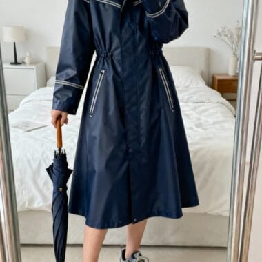 A person in a long navy raincoat and sneakers stands indoors, holding a closed umbrella, taking a mirror selfie in a bedroom.
