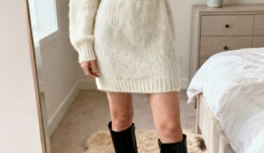 Person wearing a cream-colored knit sweater dress with a black belt and knee-high black boots, standing on a beige rug in a bedroom, taking a mirror selfie.