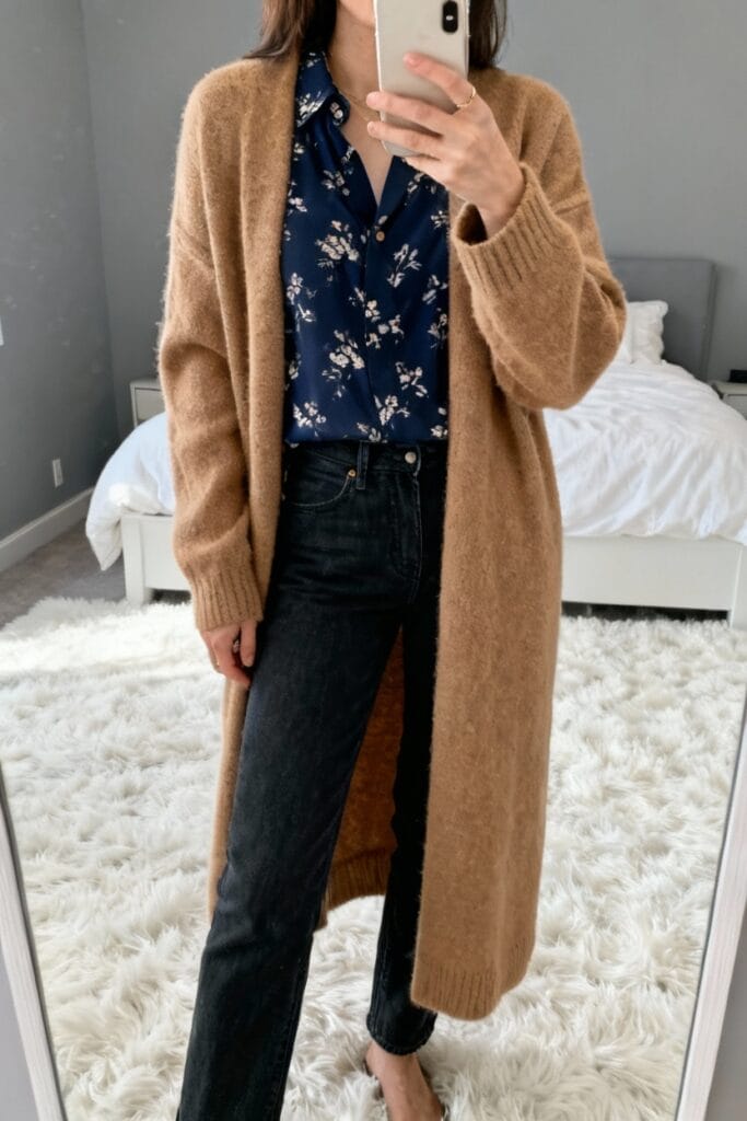Person taking a mirror selfie wearing a long camel cardigan over a blue floral blouse and black jeans, standing on a white shag rug in a bedroom.