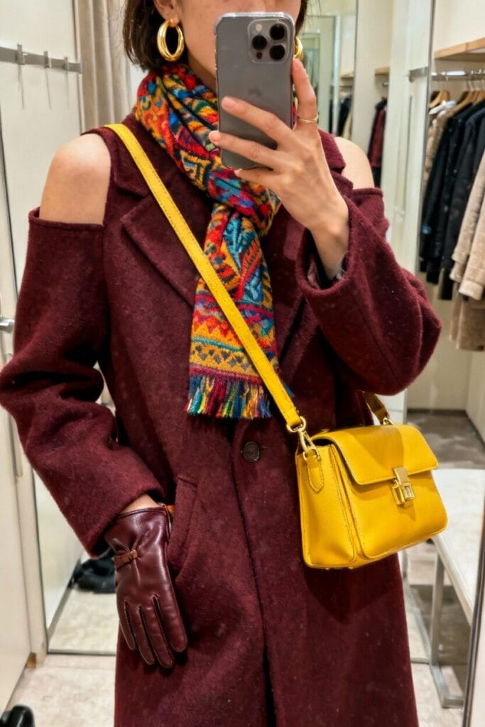 Person taking a mirror selfie wearing a burgundy coat, patterned scarf, yellow shoulder bag, gold hoop earrings, and burgundy gloves in a clothing store fitting room.