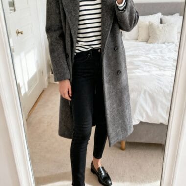 Person taking a mirror selfie wearing a gray coat, black jeans, black loafers, and a white and black striped shirt in a bedroom.