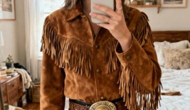 A person wearing a brown suede fringe jacket, black pants, a wide belt with a large buckle, and a brown felt hat, taking a mirror selfie in a bedroom.
